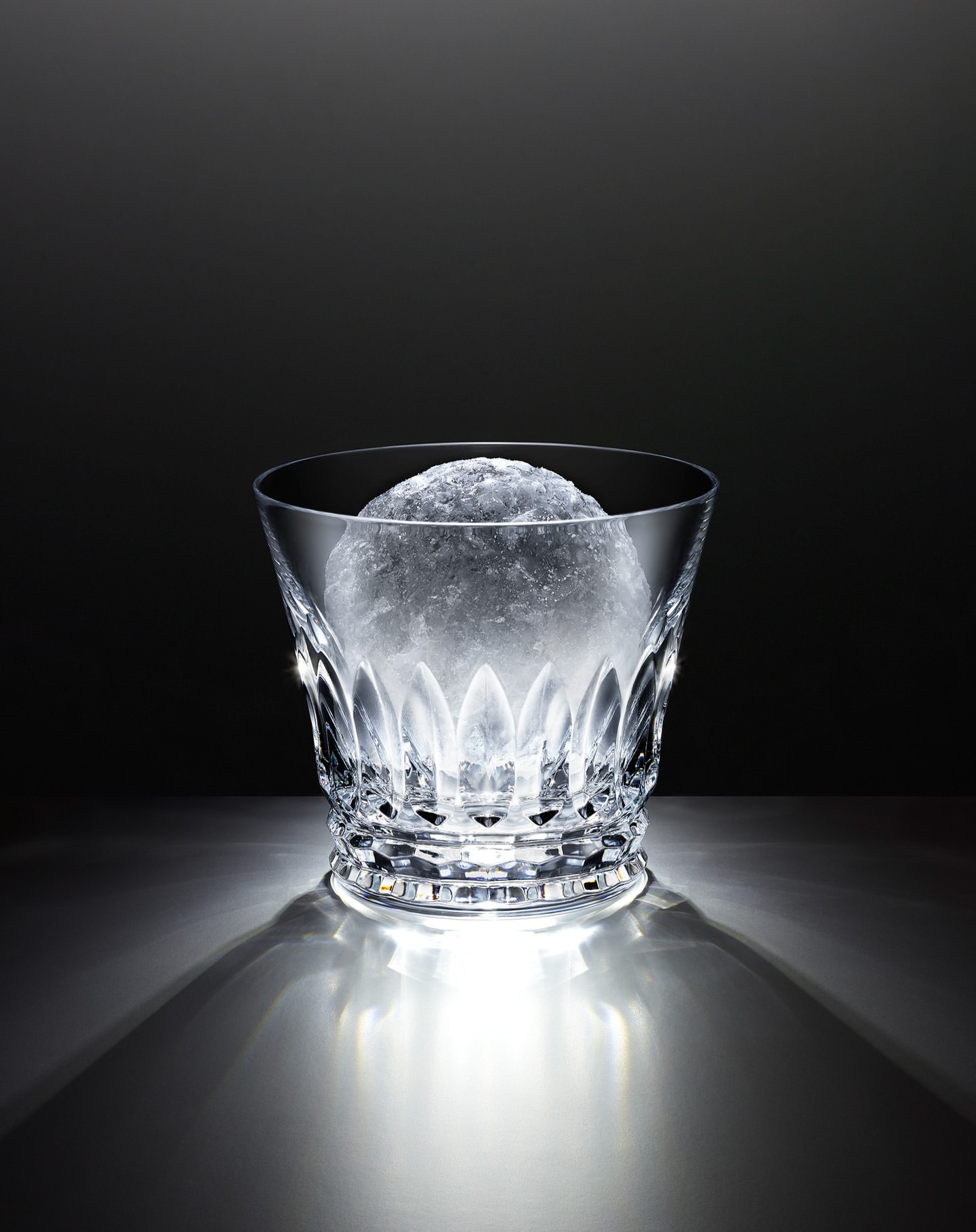 Crystal glass with faceted design, filled with crushed ice, against a dark backdrop and illuminated from below, casting shadows.