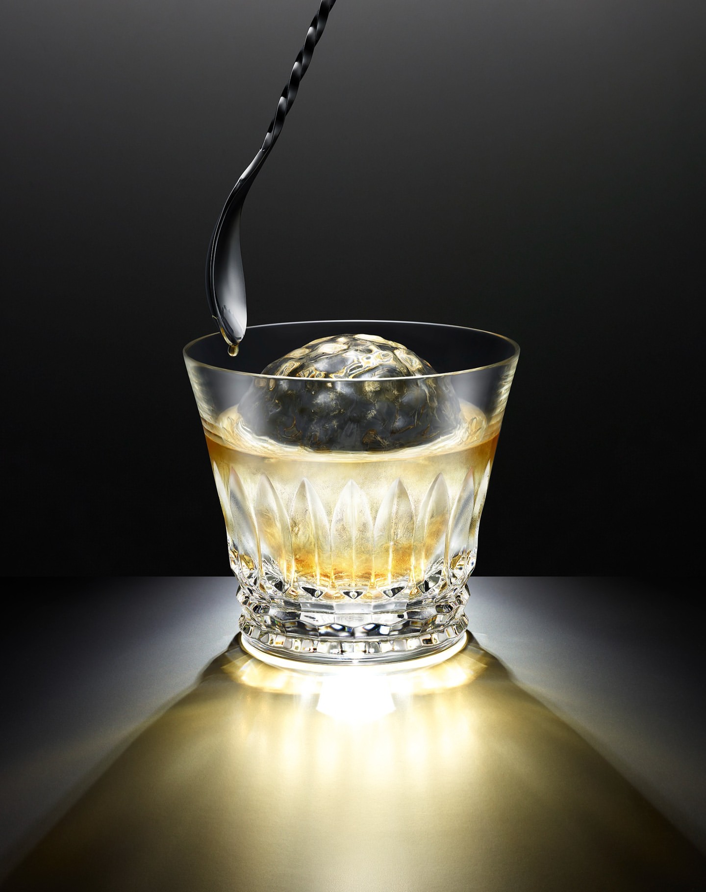 A crystal glass with a spherical ice cube and a gold liquid, with a metal spoon dripping into the beverage against a dark background.