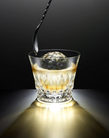 A crystal glass with a spherical ice cube and a gold liquid, with a metal spoon dripping into the beverage against a dark background.