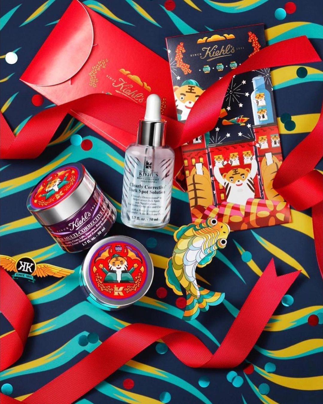 Still life of Kiehl's skincare products and red ribbon and envelope on a blue patterned background with fish decoration and confetti.