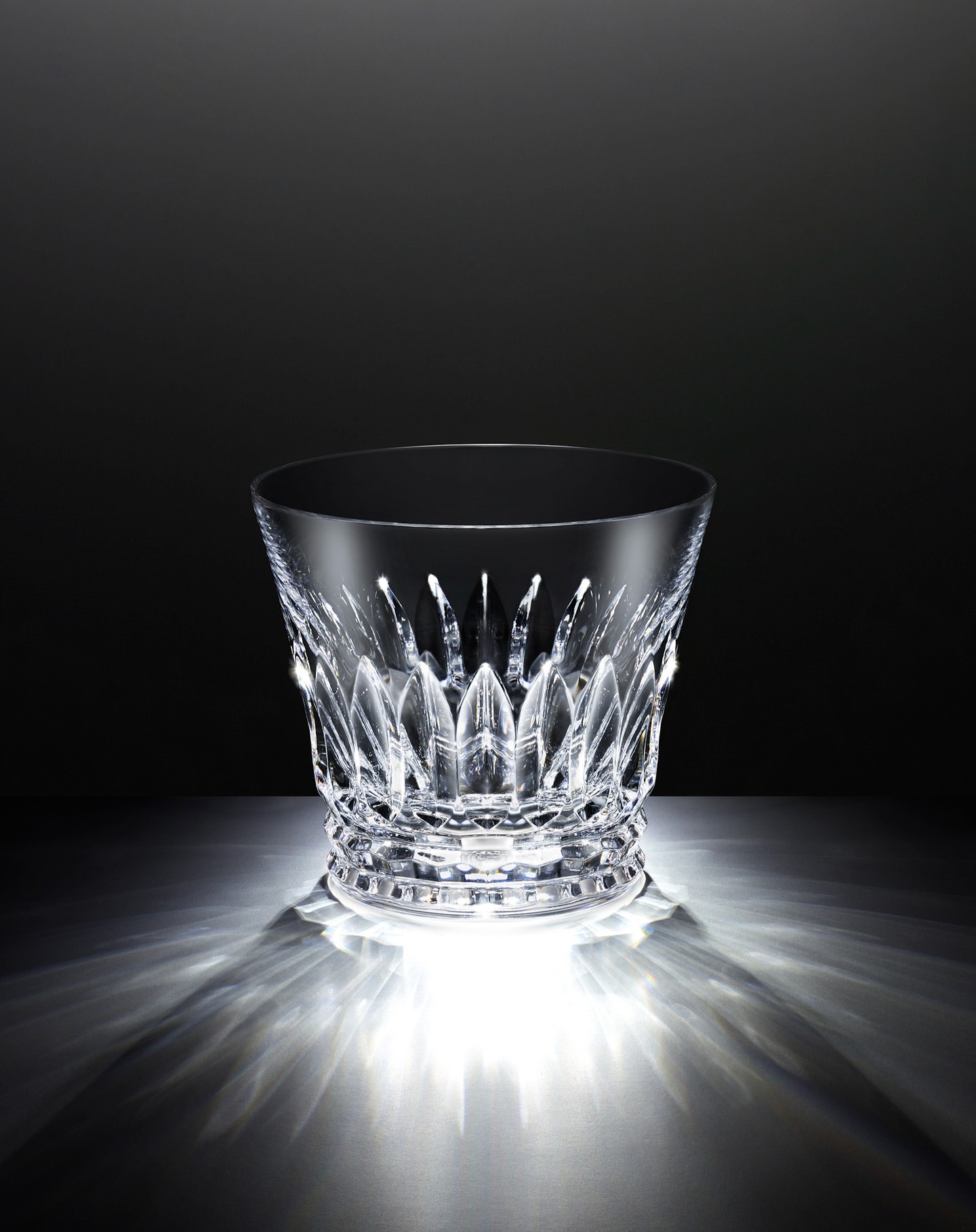 Elegant crystal ice bucket with intricate cut glass design, illuminated by bright lighting against a gradient black background.