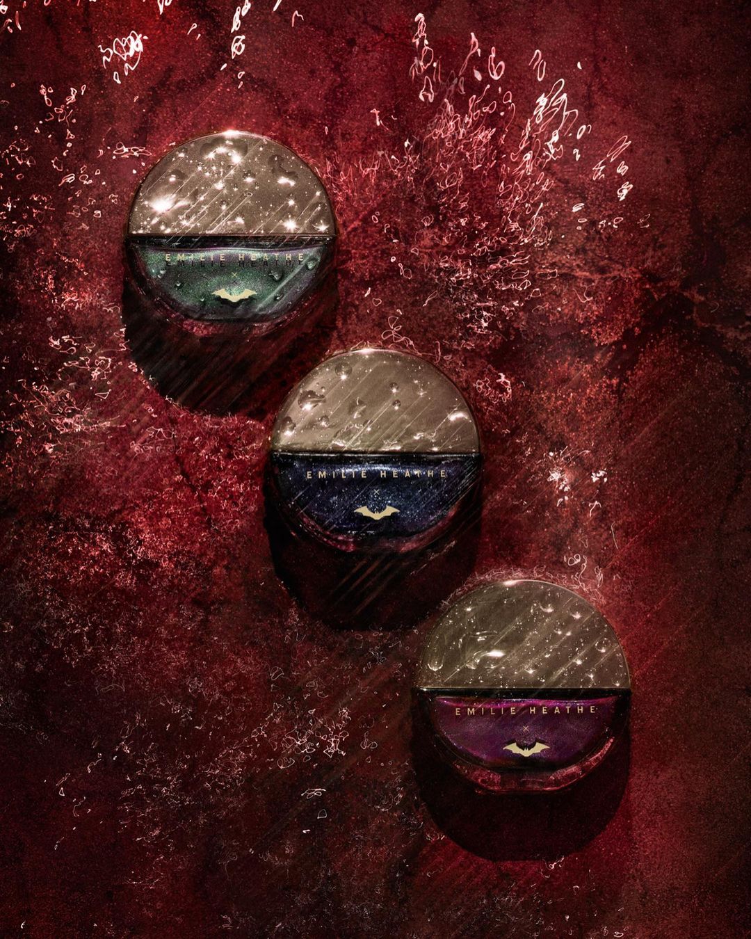 Three Emile Heathe Refillable Circle Compacts are displayed against a dark, textured red background in a beauty still life.