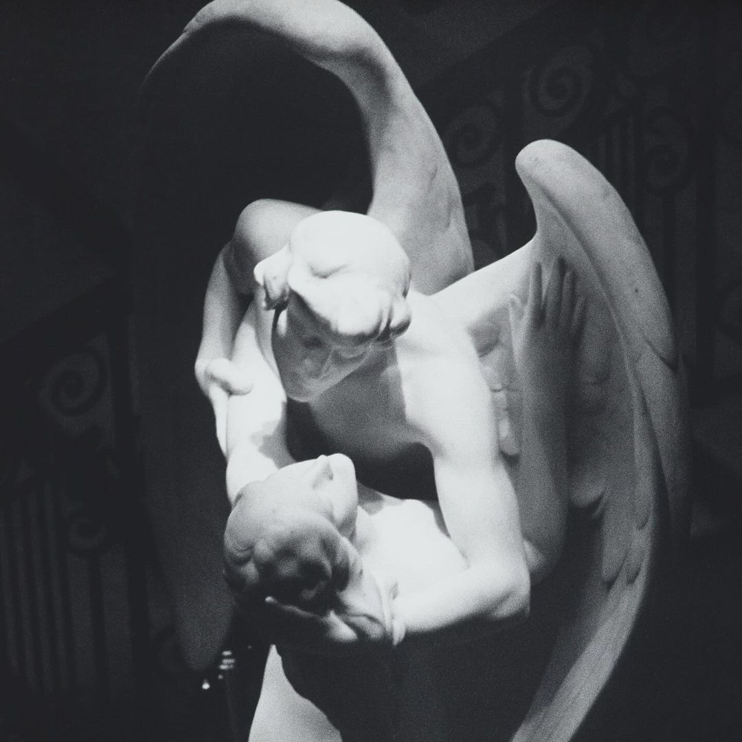 Monochrome photo of a marble sculpture depicting a winged angel holding a figure, set against a patterned background and soft lighting.