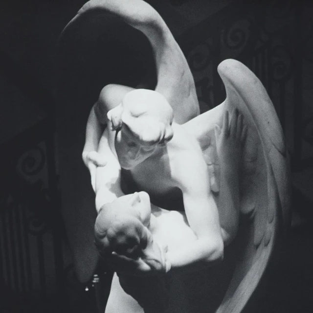 Monochrome photo of a marble sculpture depicting a winged angel holding a figure, set against a patterned background and soft lighting.