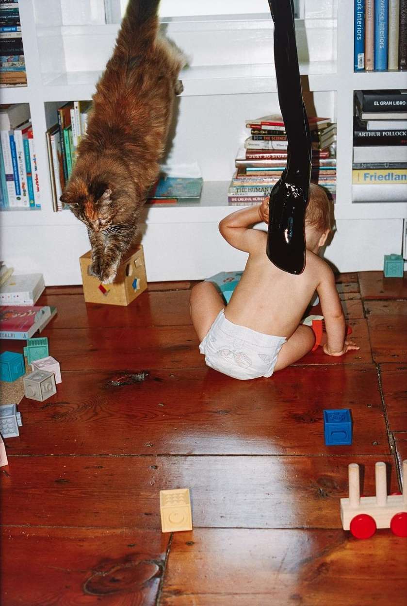 Calico cat on a toy, looking down at a baby wearing a diaper, playing with blocks on a wood floor next to a bookshelf in a home.