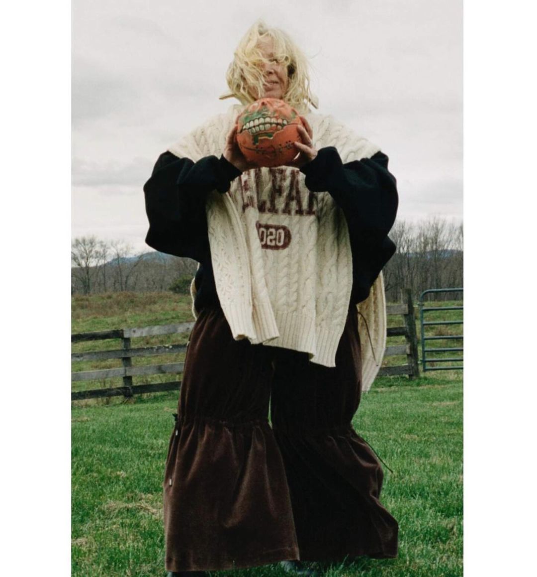 Adult female holds a painted basketball, posing outdoors in an oversized cable-knit sweater and brown ruffled pants in a grassy field.