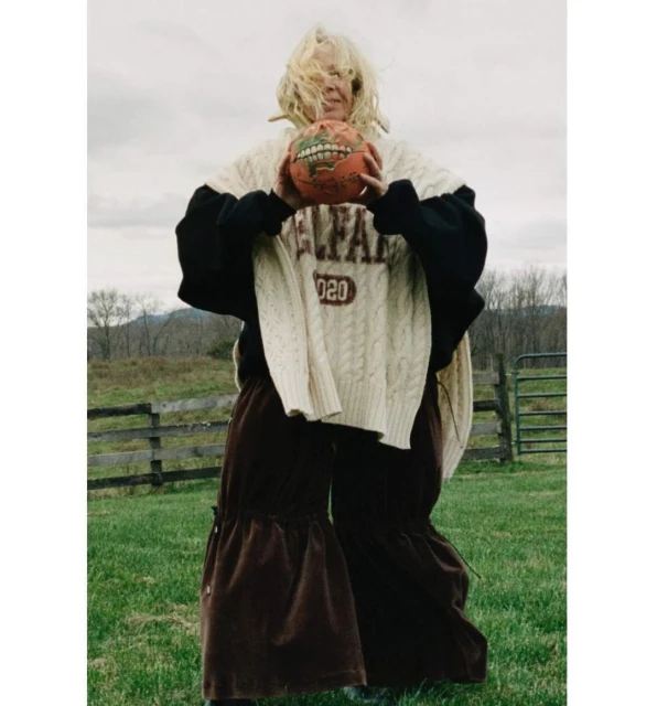 Adult female holds a painted basketball, posing outdoors in an oversized cable-knit sweater and brown ruffled pants in a grassy field.