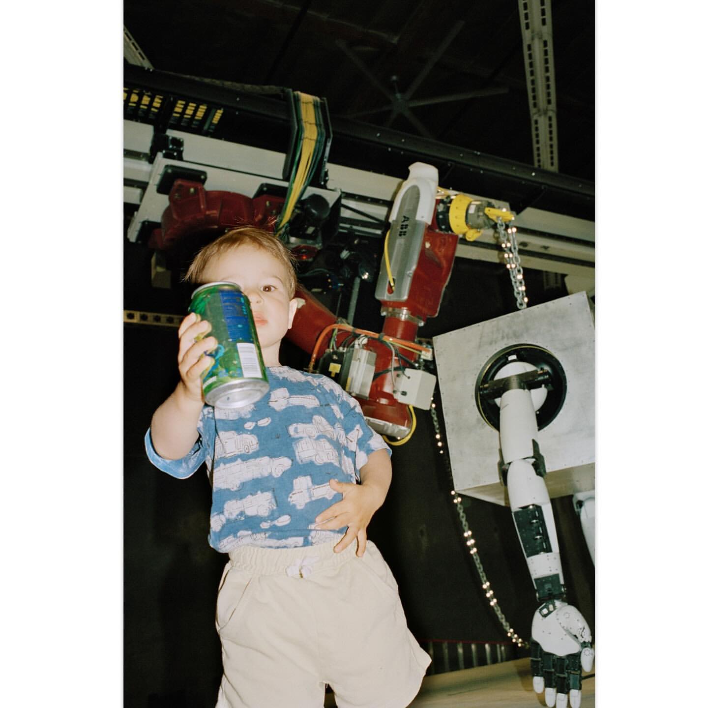 A young child in a patterned shirt holding a beverage can, positioned in front of a red and grey industrial robot in a factory setting.