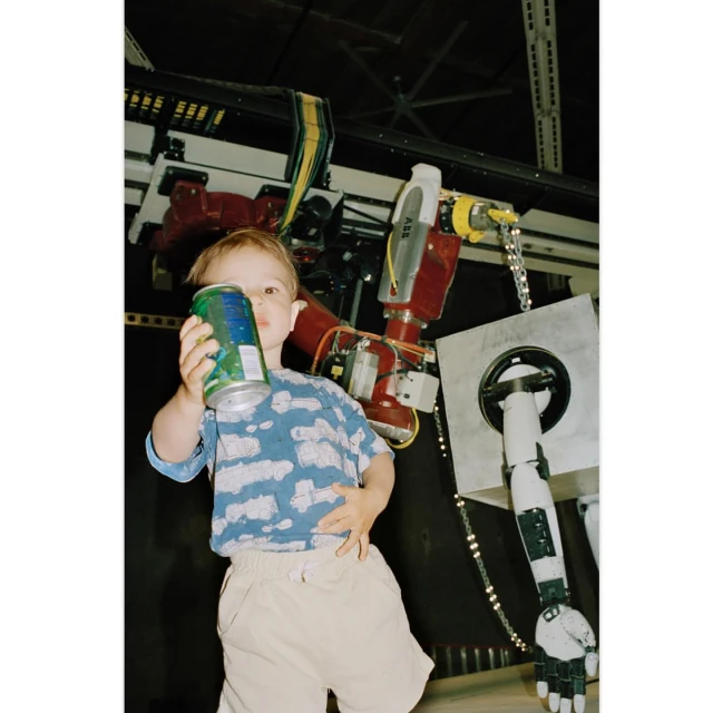 A young child in a patterned shirt holding a beverage can, positioned in front of a red and grey industrial robot in a factory setting.