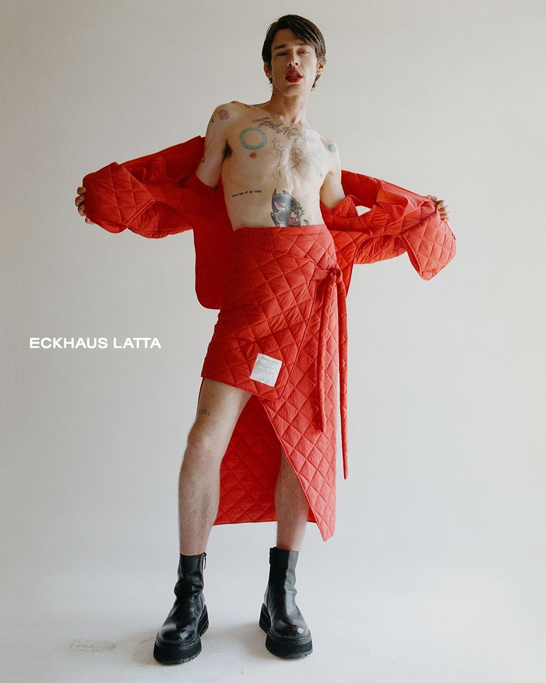 A slender adult male model poses in a studio, wearing an open red quilted jacket and matching wrap skirt with black platform boots.