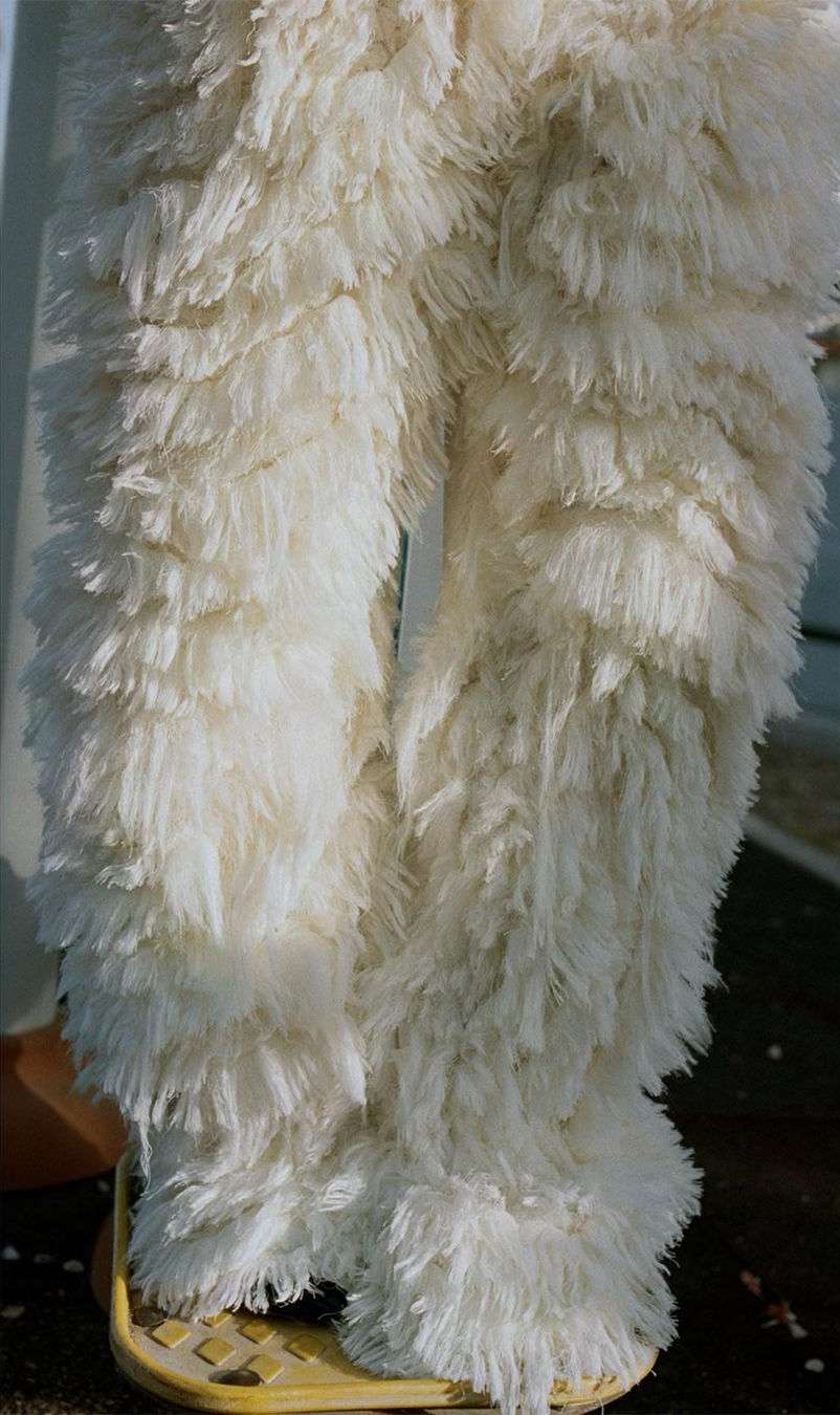 Close-up of the lower half of a person wearing shaggy, off-white faux fur pants and standing on a yellow platform, indoors.