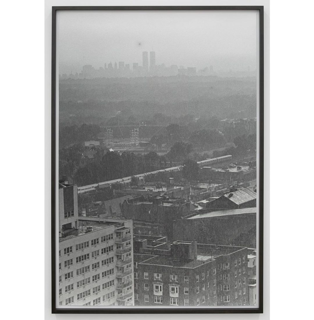 Monochromatic photograph of a cityscape with tall buildings in the foreground and skyscrapers in the distant background, framed in black.