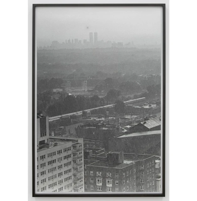 Monochromatic photograph of a cityscape with tall buildings in the foreground and skyscrapers in the distant background, framed in black.