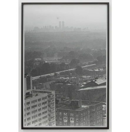 Monochromatic photograph of a cityscape with tall buildings in the foreground and skyscrapers in the distant background, framed in black.