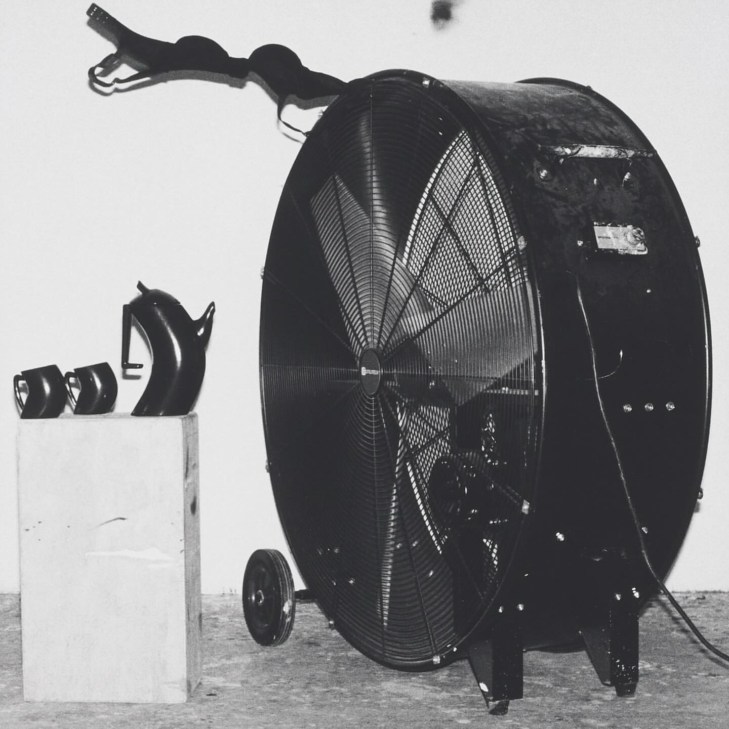 Monochromatic composition featuring a large industrial fan beside black household items atop a white block against a pale backdrop.