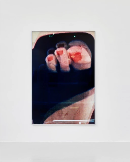 Photograph of framed foot art print on white wall with gray floor in an art gallery. The artwork has red painted toenails.