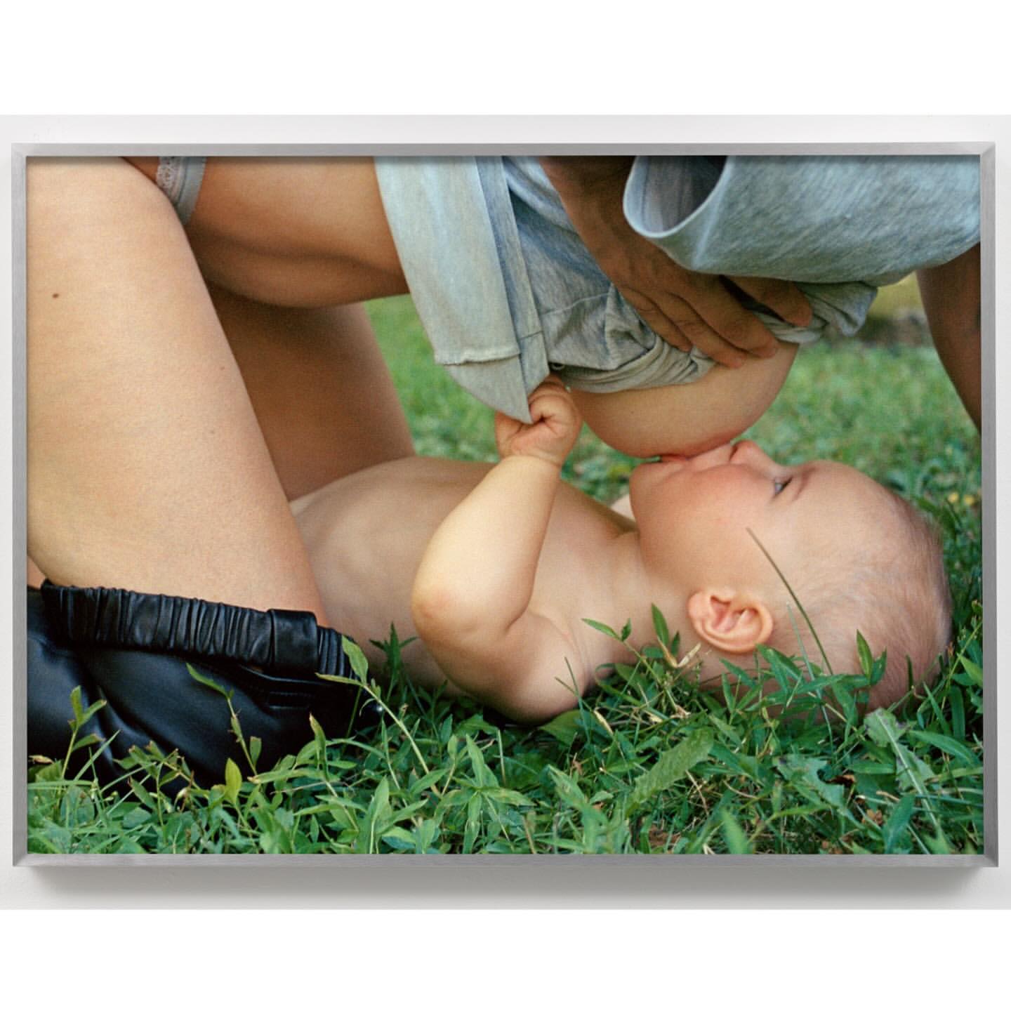 An infant is breastfed outdoors, lying on a lawn of green grass. A fair-skinned adult is positioned overhead with an off-white shirt.