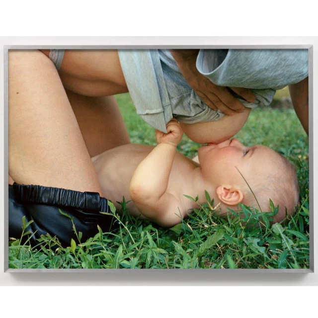 An infant is breastfed outdoors, lying on a lawn of green grass. A fair-skinned adult is positioned overhead with an off-white shirt.