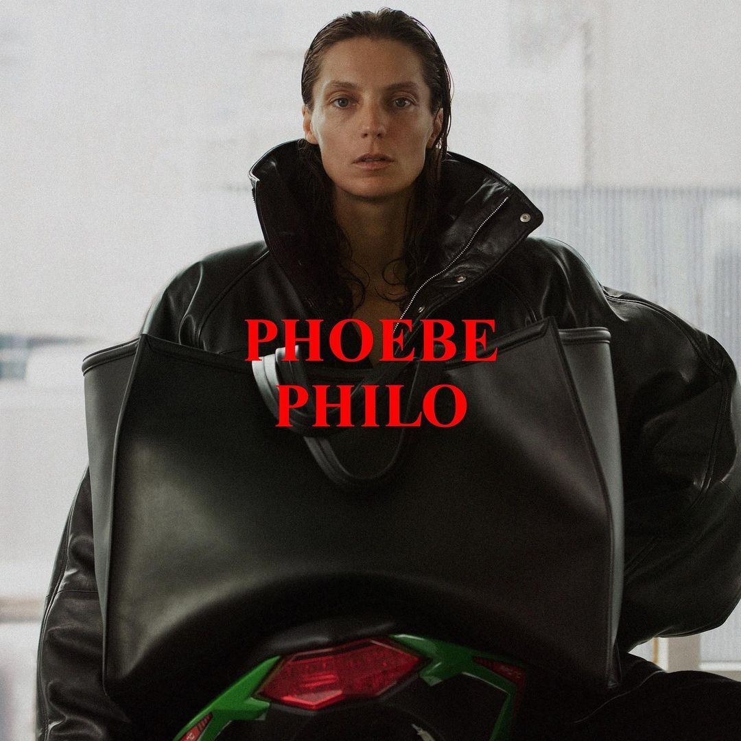 Woman with straight brunette hair wears a black leather jacket and carries a large black handbag in an indoor setting, facing the camera.