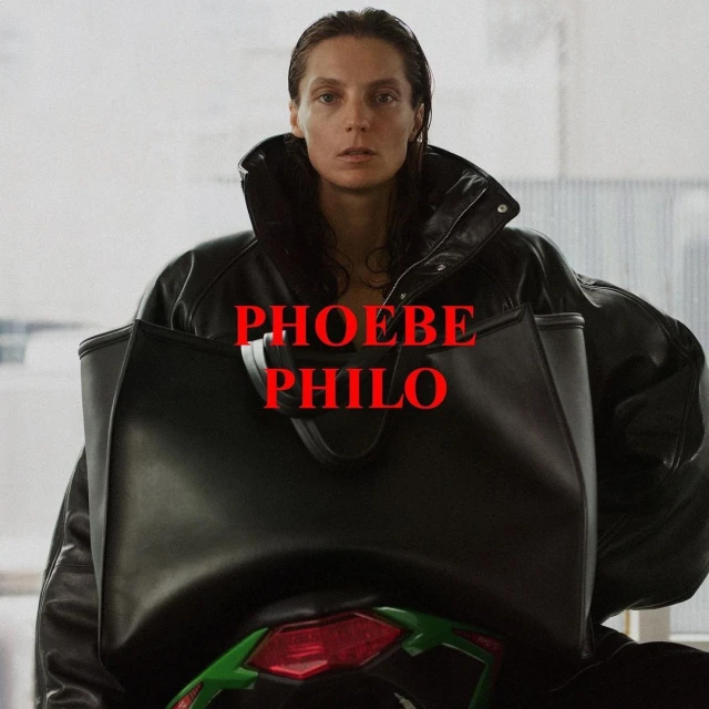 Woman with straight brunette hair wears a black leather jacket and carries a large black handbag in an indoor setting, facing the camera.