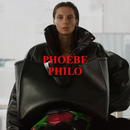 Woman with straight brunette hair wears a black leather jacket and carries a large black handbag in an indoor setting, facing the camera.