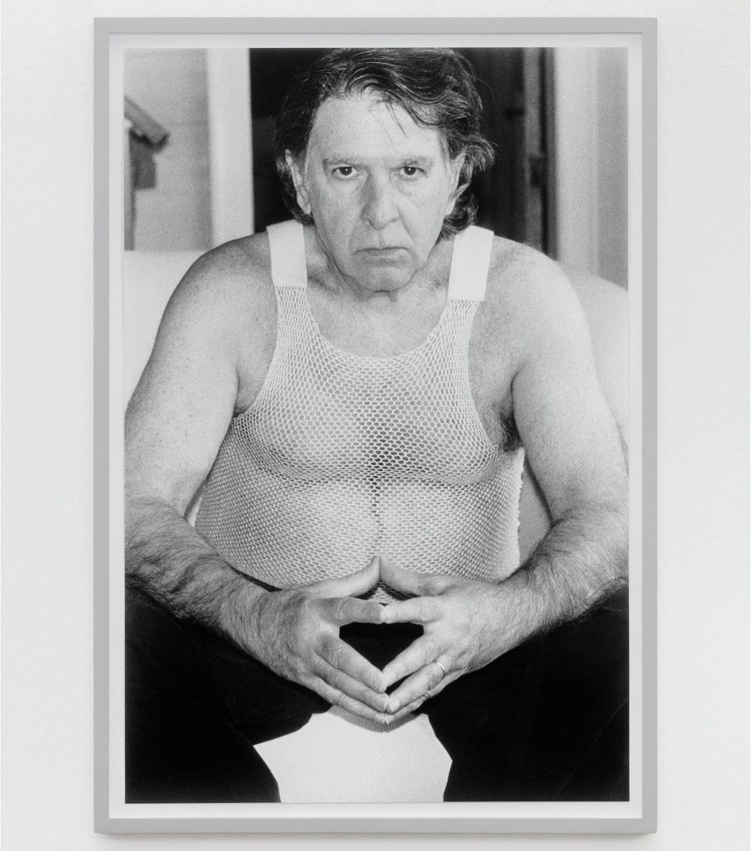 Black and white portrait of an adult man in a mesh shirt with his hands together, displayed in a gray frame on a white wall.