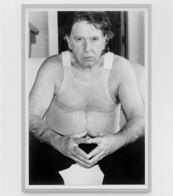 Black and white portrait of an adult man in a mesh shirt with his hands together, displayed in a gray frame on a white wall.