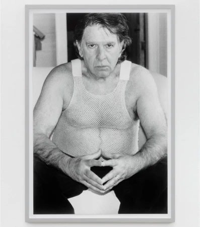 Black and white portrait of an adult man in a mesh shirt with his hands together, displayed in a gray frame on a white wall.