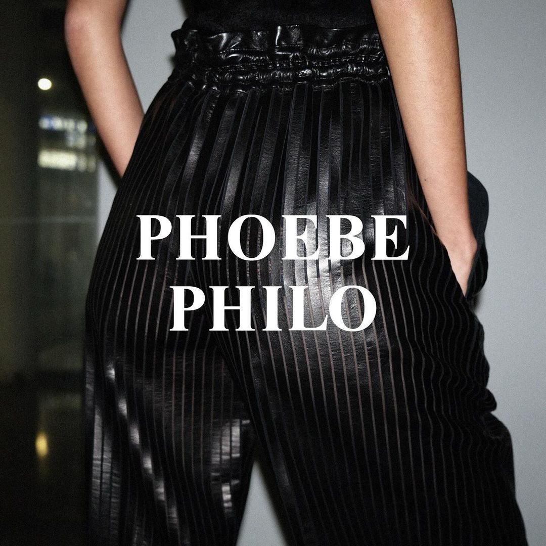 An adult female model showcases a pair of vertically striped dark, pleated pants, standing indoors against a light grey background.