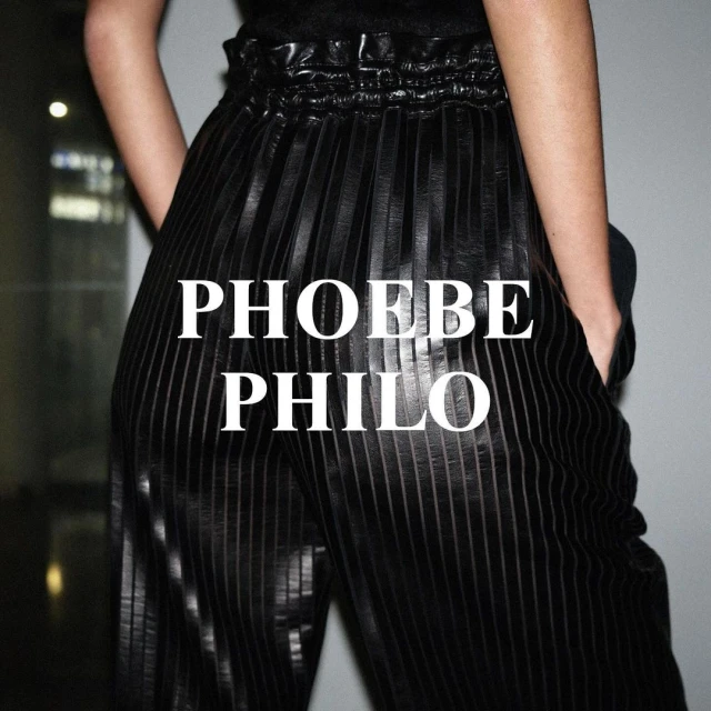 An adult female model showcases a pair of vertically striped dark, pleated pants, standing indoors against a light grey background.