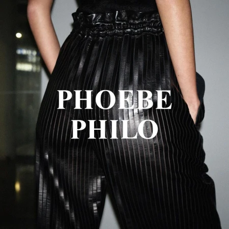 An adult female model showcases a pair of vertically striped dark, pleated pants, standing indoors against a light grey background.