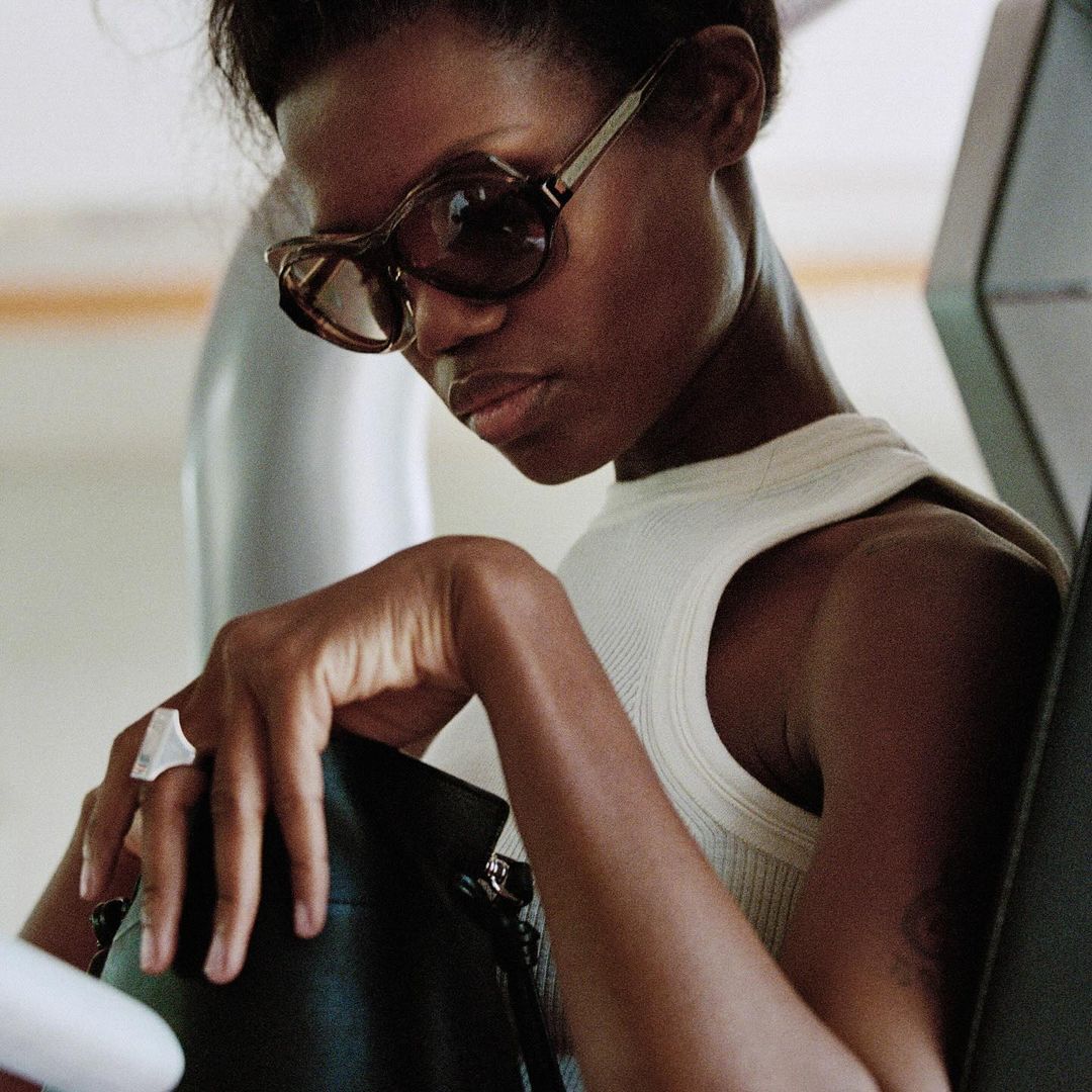 A stylish woman with sunglasses, wearing a cream top and ring, poses with a black purse in an indoor setting. Close-up shot.