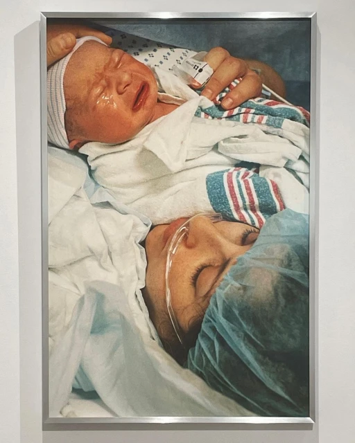 Newborn baby crying in a hospital bed with its mother. The mother is lying down with her eyes closed near her child.