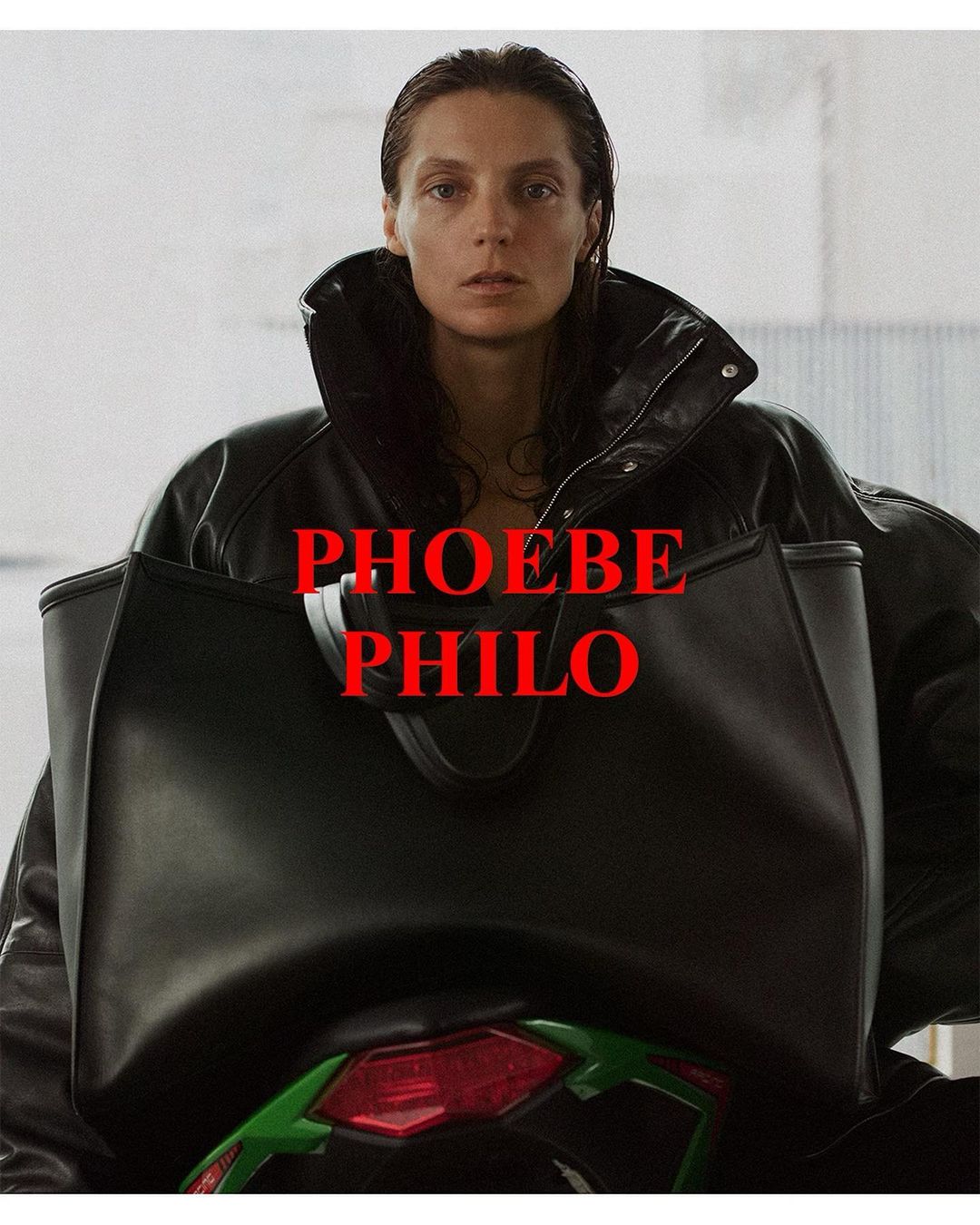 A woman with dark hair, wearing a black leather jacket and carrying a black tote bag, poses against a neutral backdrop. 'PHOEBE PHILO' is written on top.