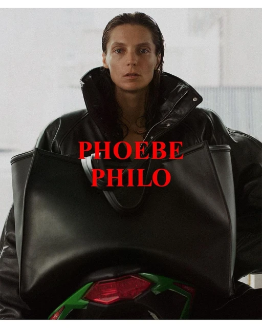 A woman with dark hair, wearing a black leather jacket and carrying a black tote bag, poses against a neutral backdrop. 'PHOEBE PHILO' is written on top.