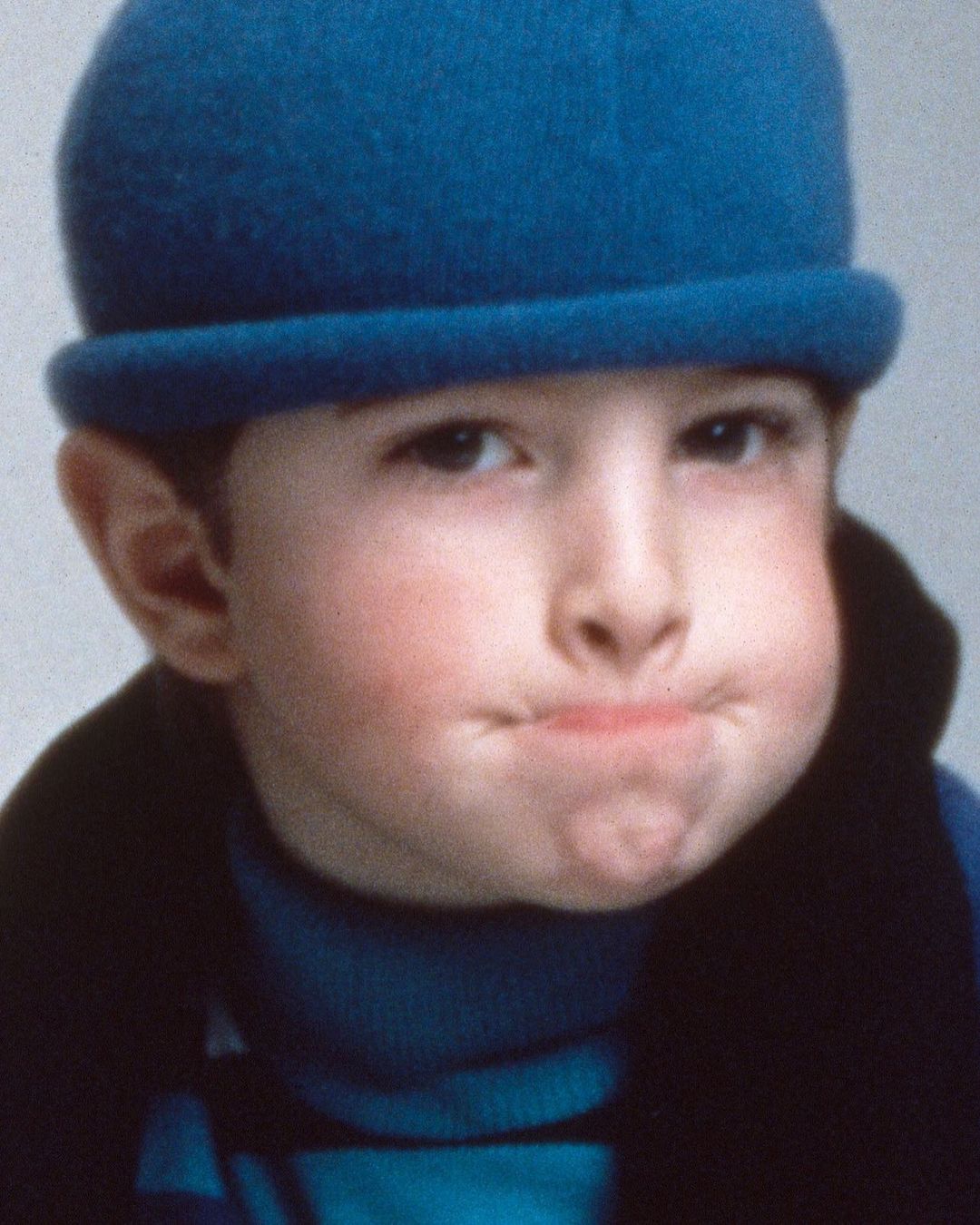 Young child portrait. Close-up of a kid wearing a blue hat and turtleneck sweater, with puffed cheeks, against a neutral backdrop.