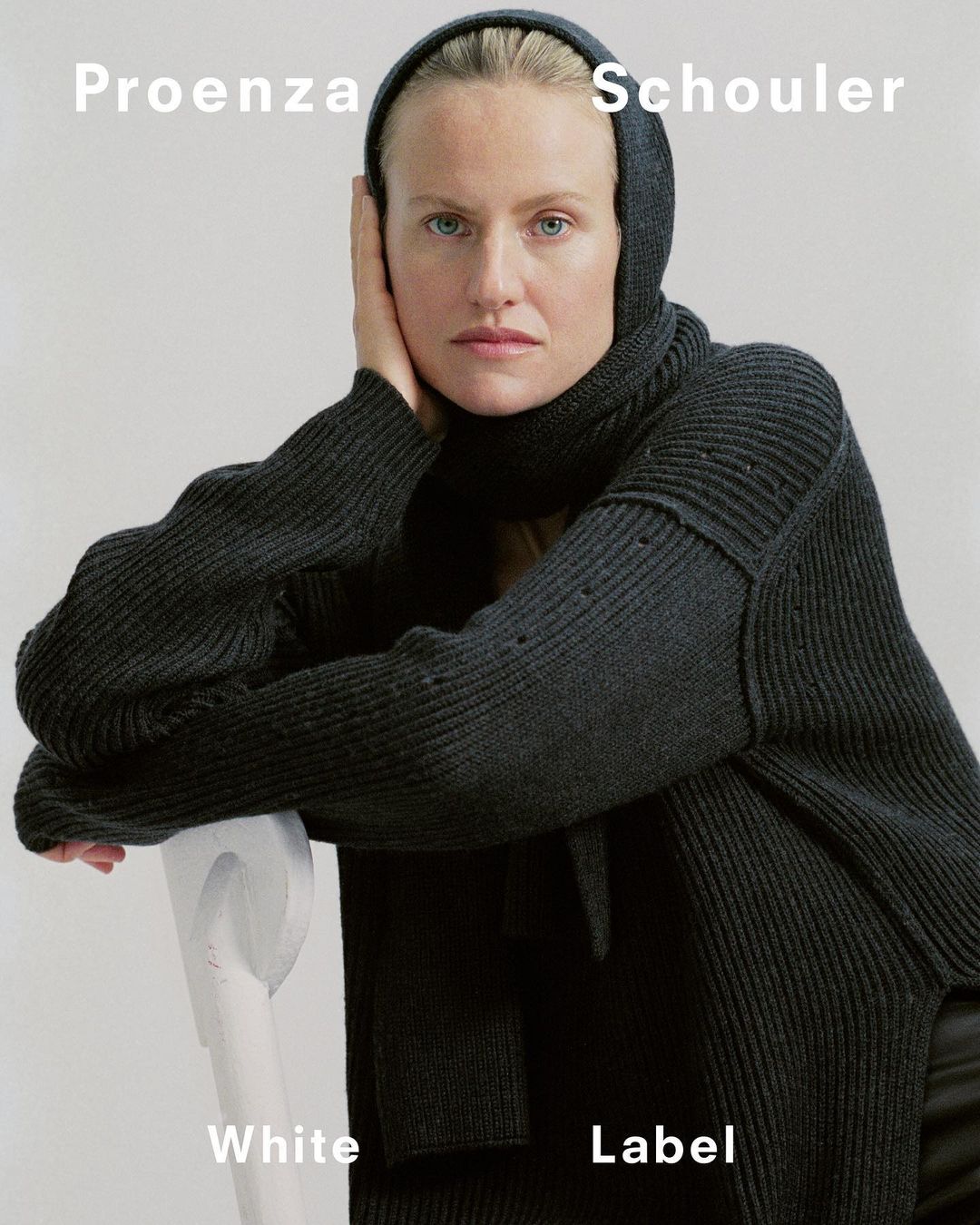 Fashion portrait of a fair-skinned woman wearing a dark grey knit sweater and matching hood, shot in a bright indoor setting.