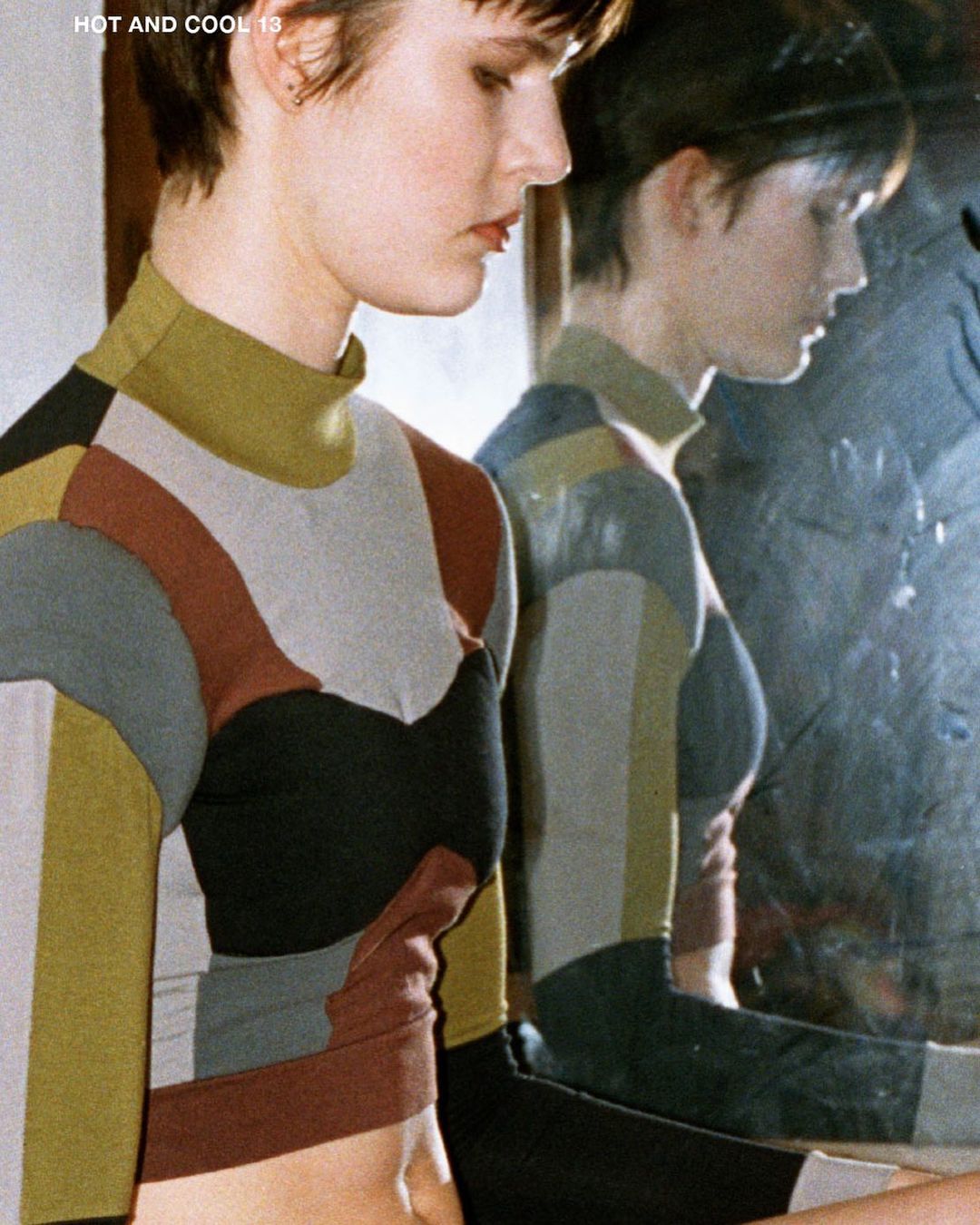 Young woman with short hair in a color block long-sleeve shirt, posing next to her reflection in a distorted mirror. Fashion style.