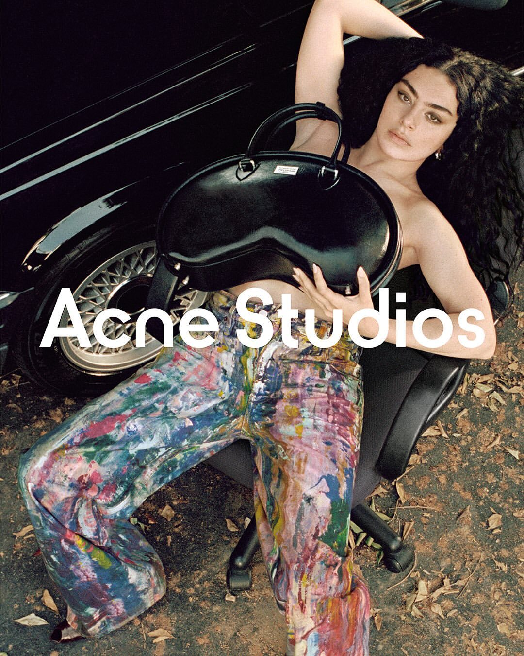 Brunette woman with a black purse sitting on an office chair next to a black car while wearing paint-splattered pants.