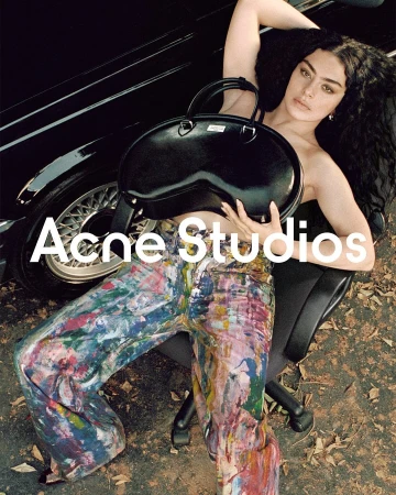 Brunette woman with a black purse sitting on an office chair next to a black car while wearing paint-splattered pants.