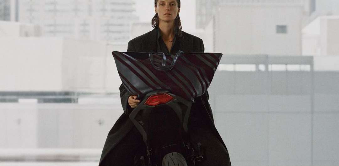 Fashion shot of a woman in a black coat on a motorcycle holding a striped tote bag in an urban environment with neutral tones.