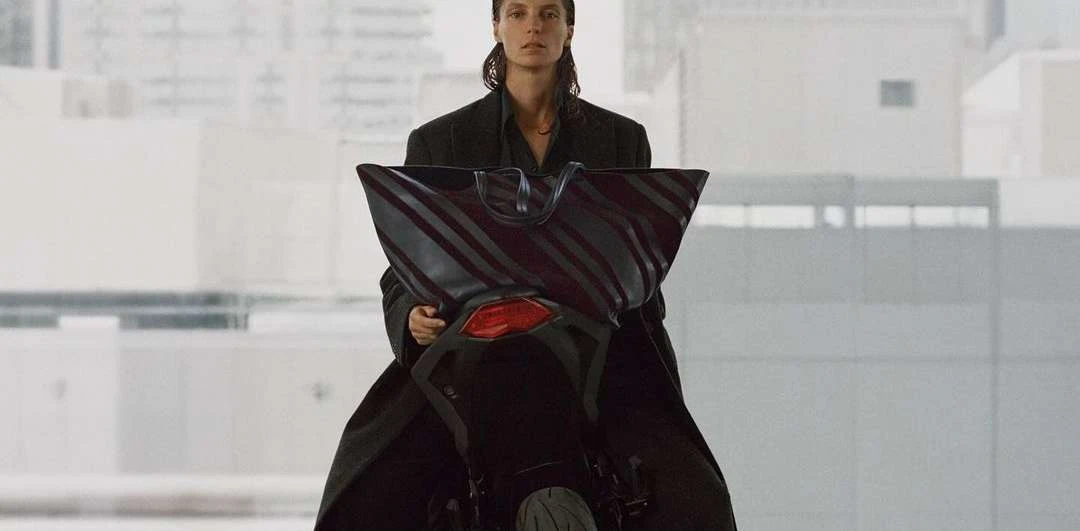 Fashion shot of a woman in a black coat on a motorcycle holding a striped tote bag in an urban environment with neutral tones.