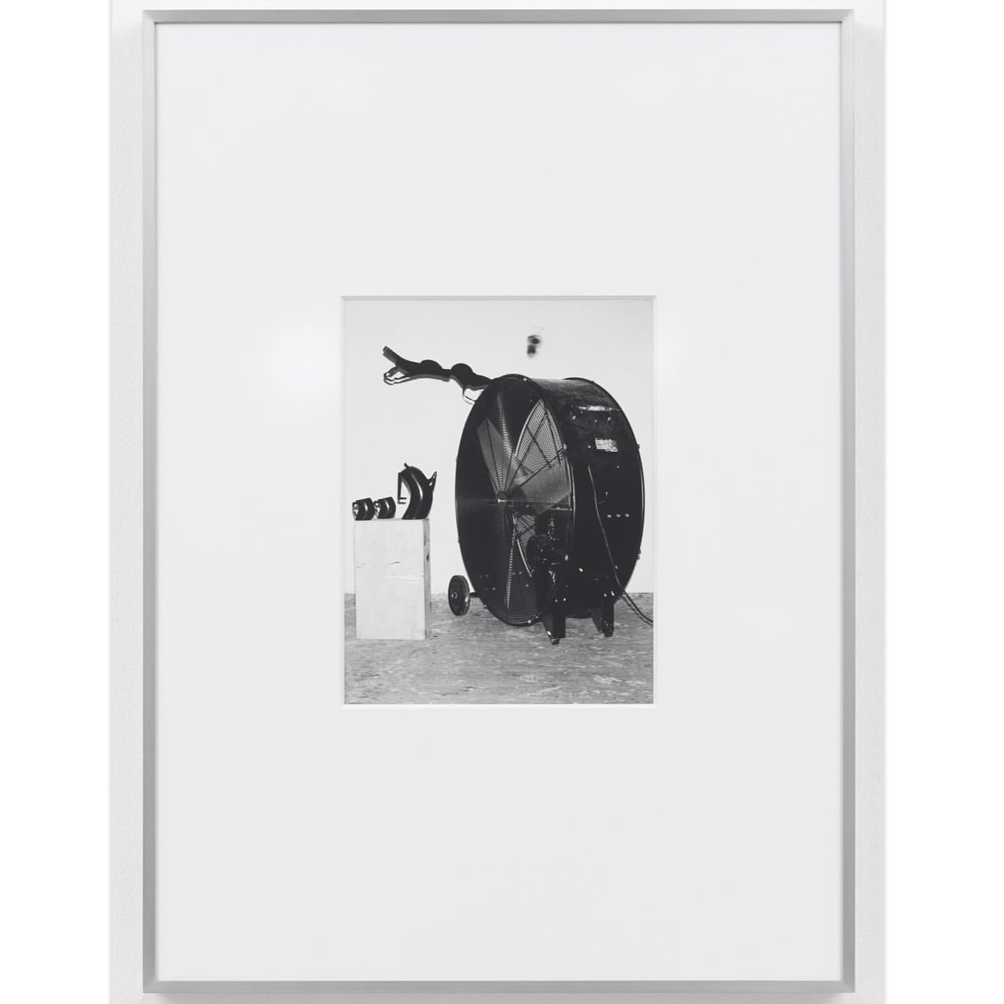 Black and white photograph of a large fan on wheels next to a box and other objects, presented in a white frame and white background.