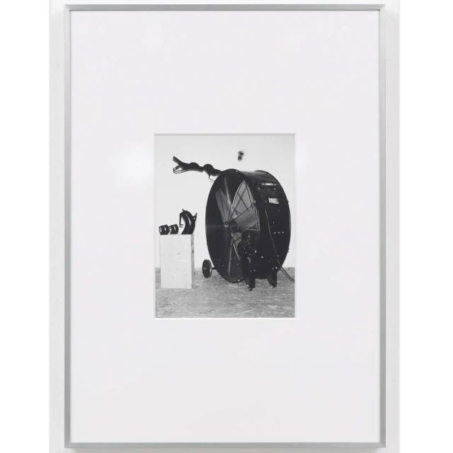 Black and white photograph of a large fan on wheels next to a box and other objects, presented in a white frame and white background.