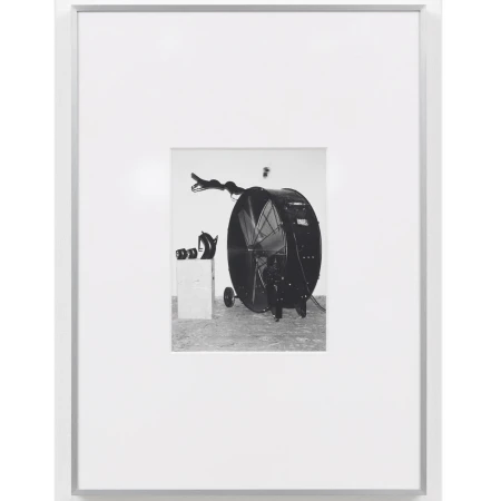 Black and white photograph of a large fan on wheels next to a box and other objects, presented in a white frame and white background.