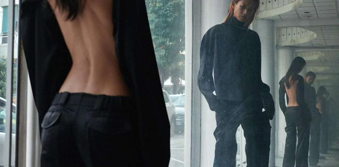 Back view of a slender woman in an open-back jacket and black trousers. Another woman with red hair poses in similar attire in a mirror.