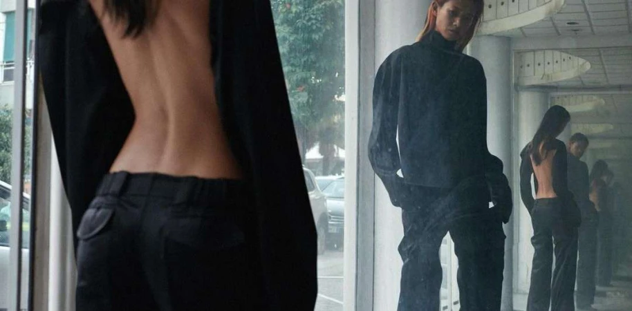 Back view of a slender woman in an open-back jacket and black trousers. Another woman with red hair poses in similar attire in a mirror.