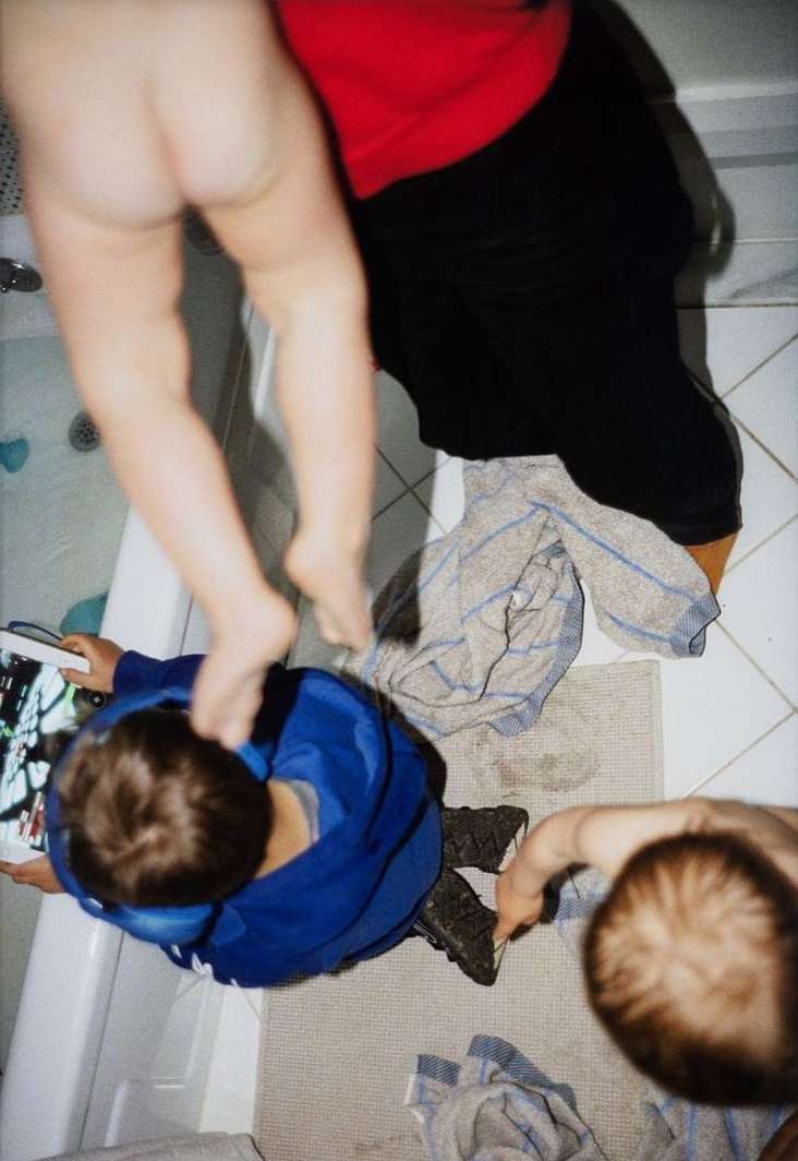 Overhead shot of two adults and a child interacting in a bathroom with tiled floors and a bathtub. One adult partially nude.