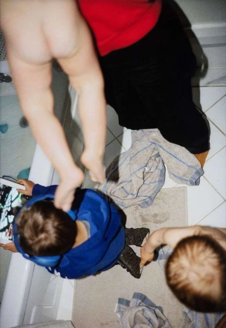 Overhead shot of two adults and a child interacting in a bathroom with tiled floors and a bathtub. One adult partially nude.