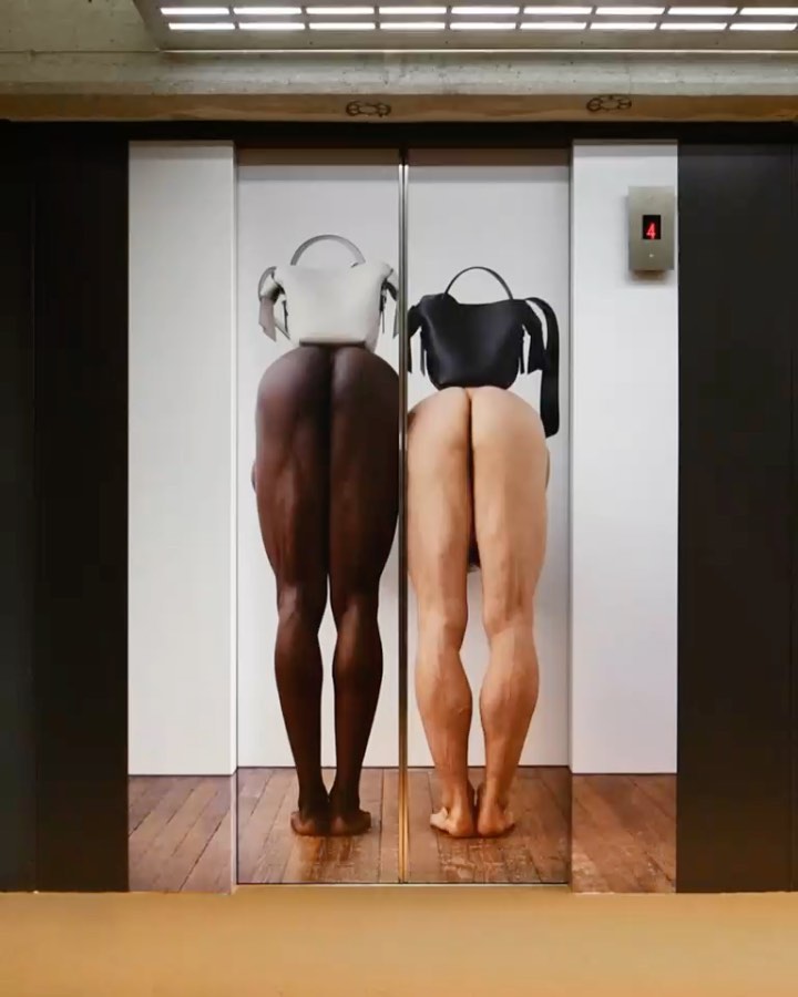 Conceptual art piece featuring two figures with skin in contrasting tones wearing handbags in an elevator with metallic and light fixtures.